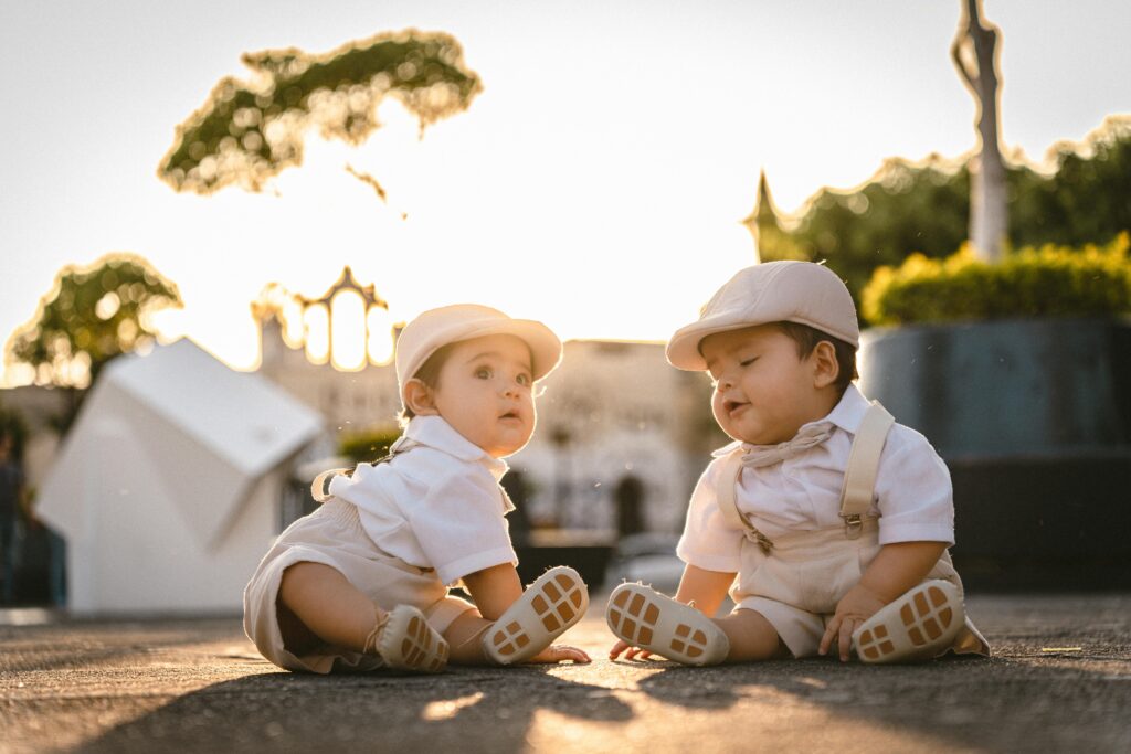 Twin babies in adorable white outfits sitting outdoors at sunset, exuding cuteness and warmth.